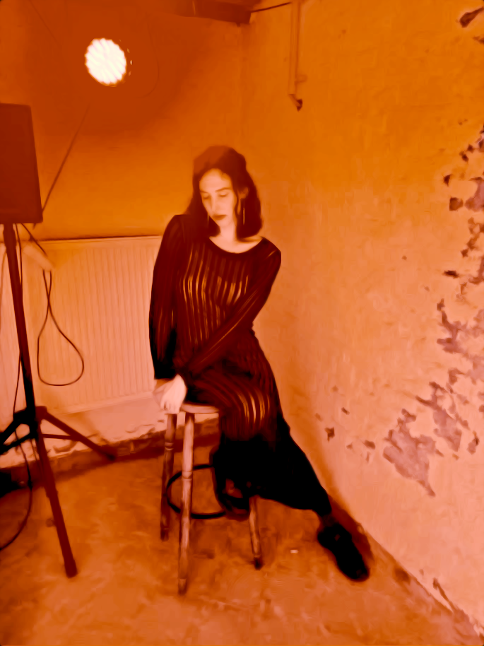 tsita zee dreamily sitting on wooden stool next to loudspeaker, warm and diffuse orange lighting.