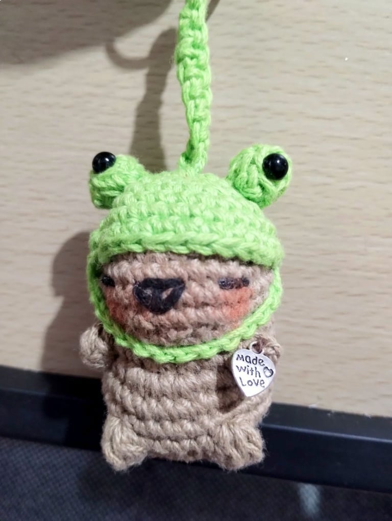 Llavero de capibara tejido a crochet, con un gorro de rana verde y un dije de corazón plateado.