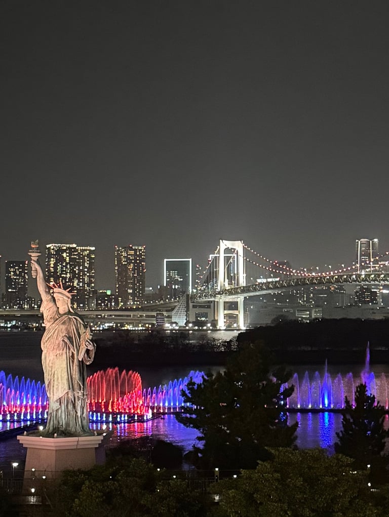 The Statue of Liberty in Odaiba, Tokyo at night, with the illuminated Rainbow Bridge and city skyline.