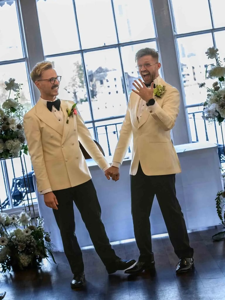 Two grooms in cream tuxedos holding hands and celebrating after their indoor wedding ceremony.