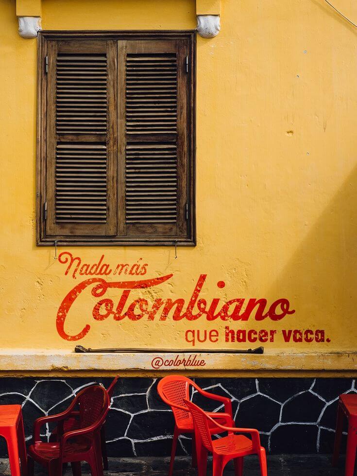Rustic wooden shutters on a yellow wall with Colombian Spanish text and red chairs.
