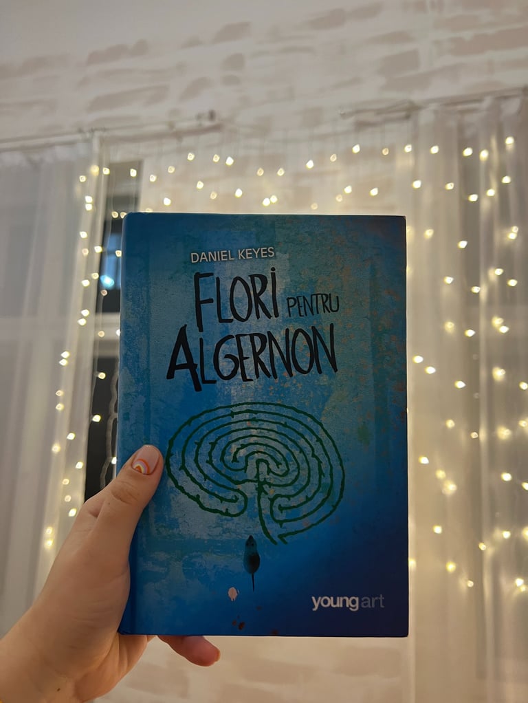 Mână ținând cartea 'Flori pentru Algernon' de Daniel Keyes pe un fundal de luminițe de basm.