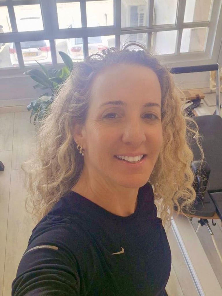Smiling woman with curly hair wearing activewear in a pilates studio with reformer equipment.