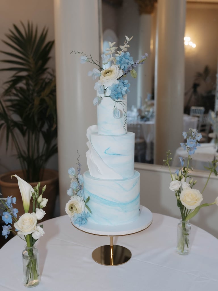 Three-tier white and blue marbled wedding cake decorated with fresh blue and white flowers on a gold stand.
