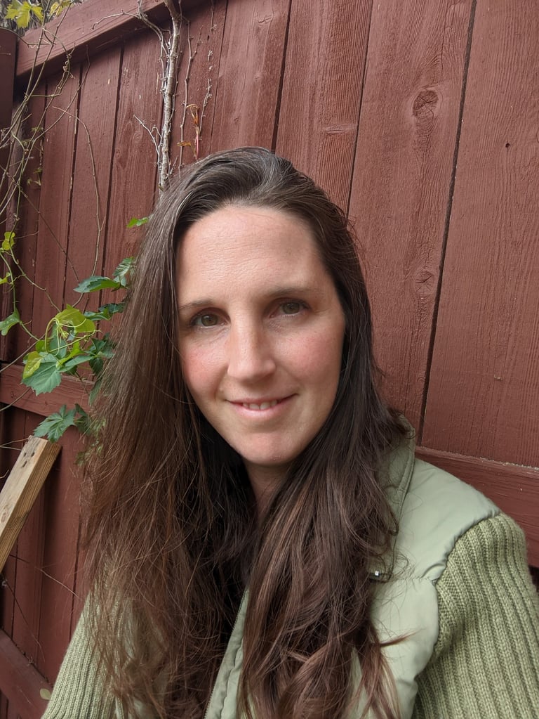 A smiling woman with long brown hair wearing a green vest against a rustic red wooden fence.