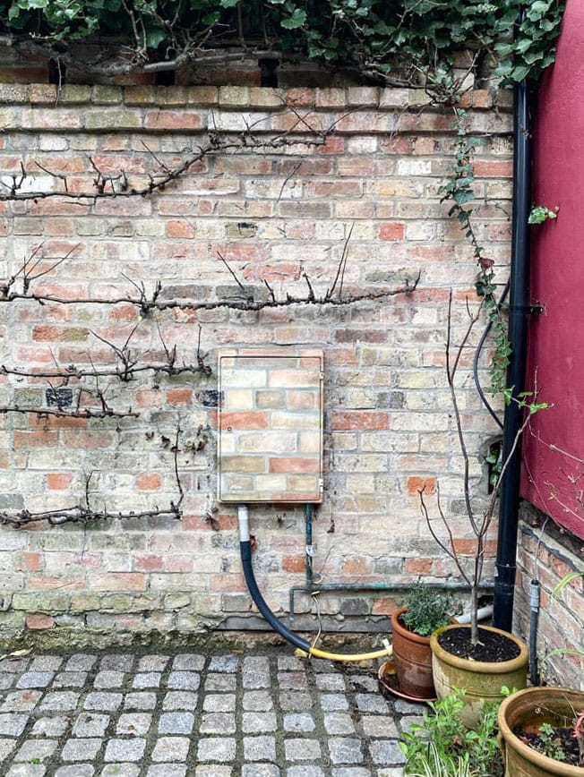 An electricity box attached to a garden brick wall painted in camouflage brick pattern.