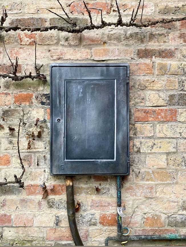 An electricity box in a garden painted to resemble an antique faux lead cartouche.