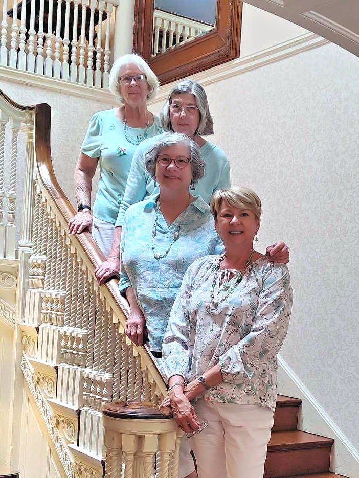 Four smiling senior women posing together on a historic wooden staircase in a bright home.
