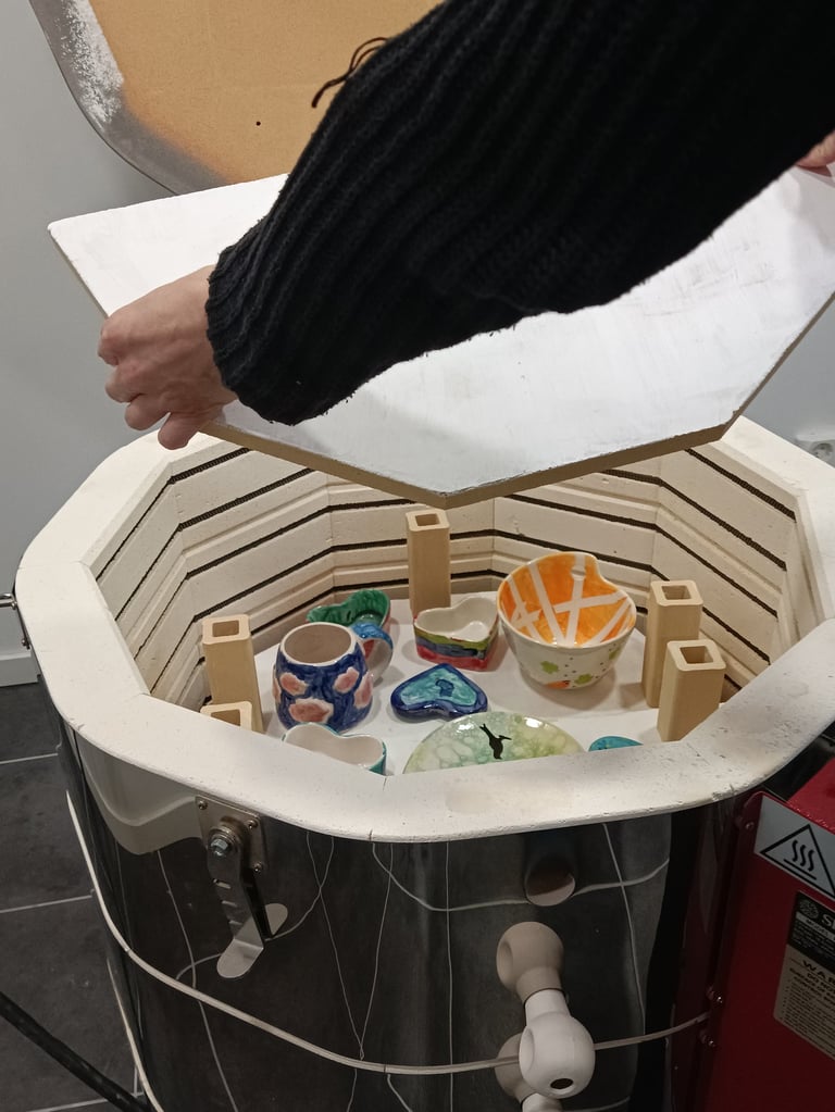 A hand opening a ceramic kiln filled with colorful glazed pottery bowls and mugs.