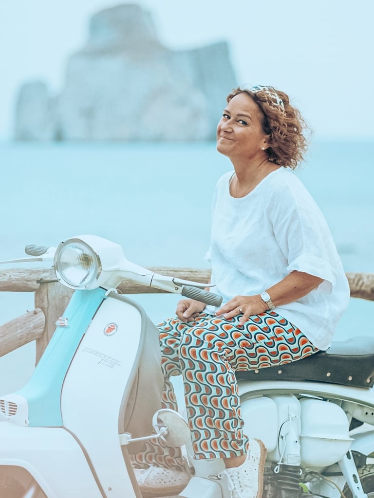 Alessandra Polo on a Vespa with view of Pan di Zucchero sea stack in Sardinia.