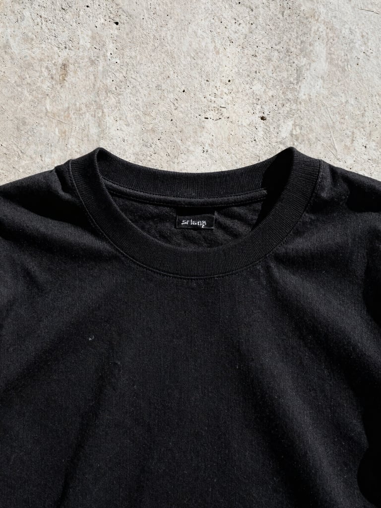 Low-angle close-up of a heavy-weight 250 GSM black cotton t-shirt showing the thick tight collar and dense fabric texture against a raw concrete Manila wall under harsh midday sunlight.