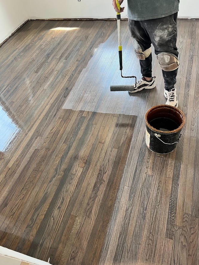 Professional applying a glossy polyurethane finish to dark stained oak hardwood floors using a paint roller.