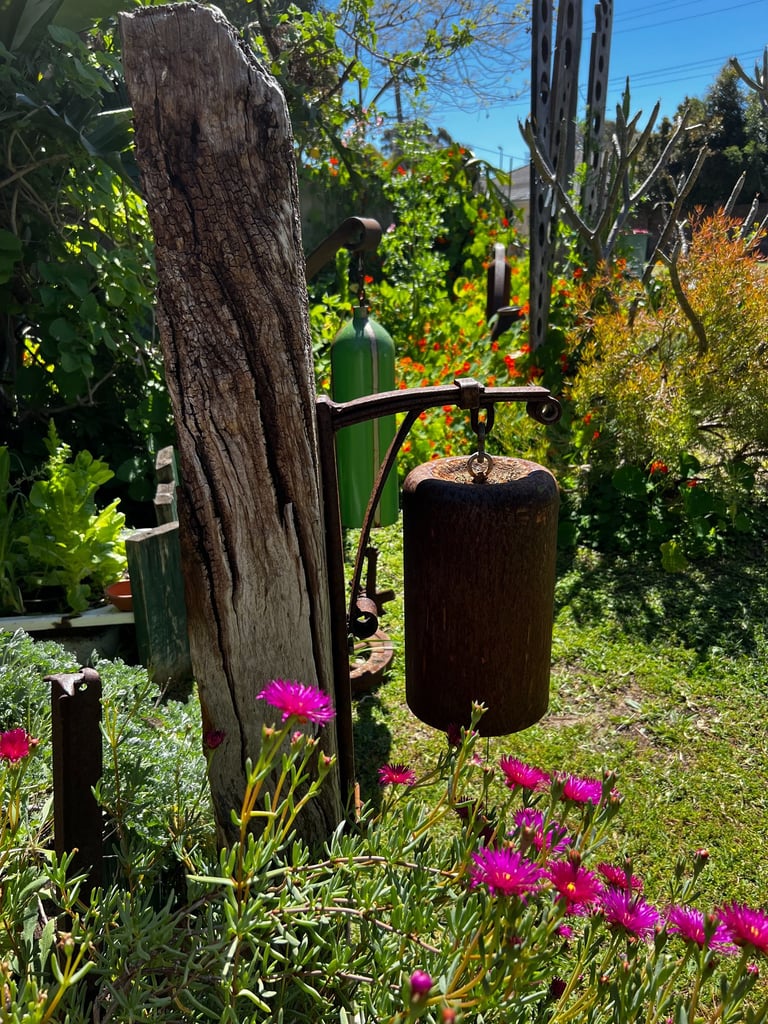 Rustic garden bells hanging from a wooden post amidst vibrant pink flowers and lush greenery.