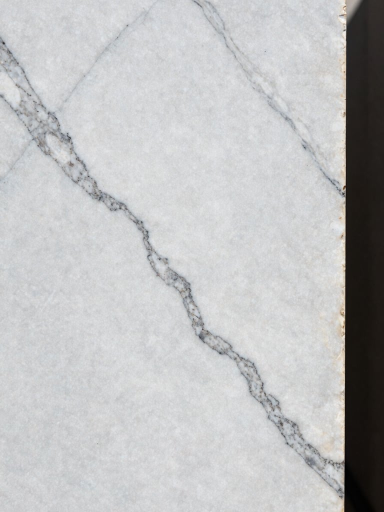 Extreme close-up of a Carrara marble slab surface in north-facing daylight, sharp white ground with deep grey veining running diagonally across the full frame, stone edge visible at the right, no staging or props