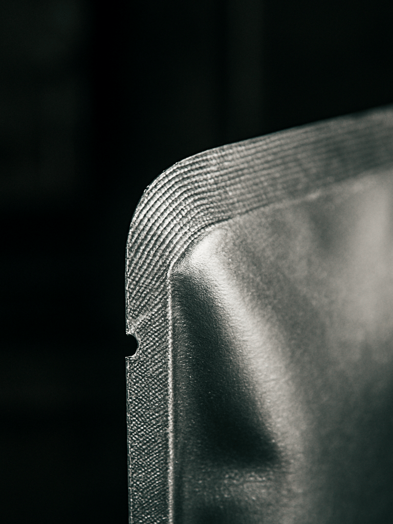 Angled studio close-up of a die-cut sachet edge being examined under manufacturing conditions, strobe lighting from the right illuminating the precise die-cut seam and foil surface texture, dark industrial background, focus on the mechanical sealing detail along the custom contour