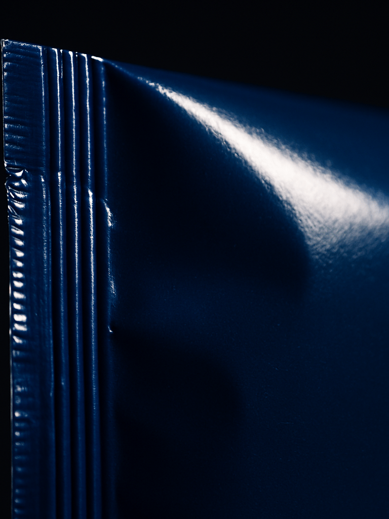 Extreme close-up of a sealed sachet edge under studio strobe lighting, the heat-seal seam razor-sharp in focus, deep blue flexible film catching hard specular light, sachet positioned to the left third of the frame with dark negative space to the right