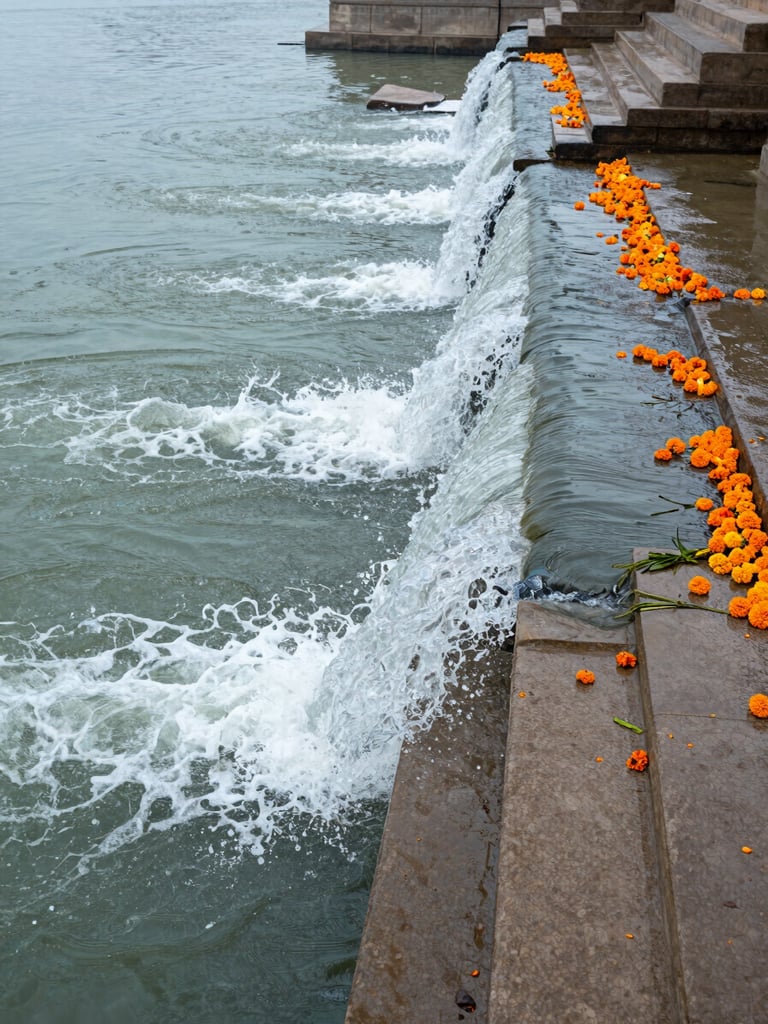 Close wide shot of the Ganga in motion, water breaking over submerged stone steps at a ghat, diffused morning daylight, pilgrims' orange marigold offerings drifting downstream on the right edge, muted blue-green water tones