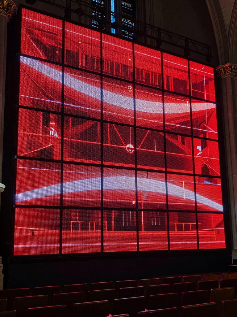 Wide environmental shot of a large installed LED wall dominating the front of a darkened church sanctuary, the screen blazing with deep red and white graphics during a live production, cinematic tungsten stage lighting raking across the room from the sides, rows of empty seats in the foreground, the wall's seamless panels glowing with controlled precision
