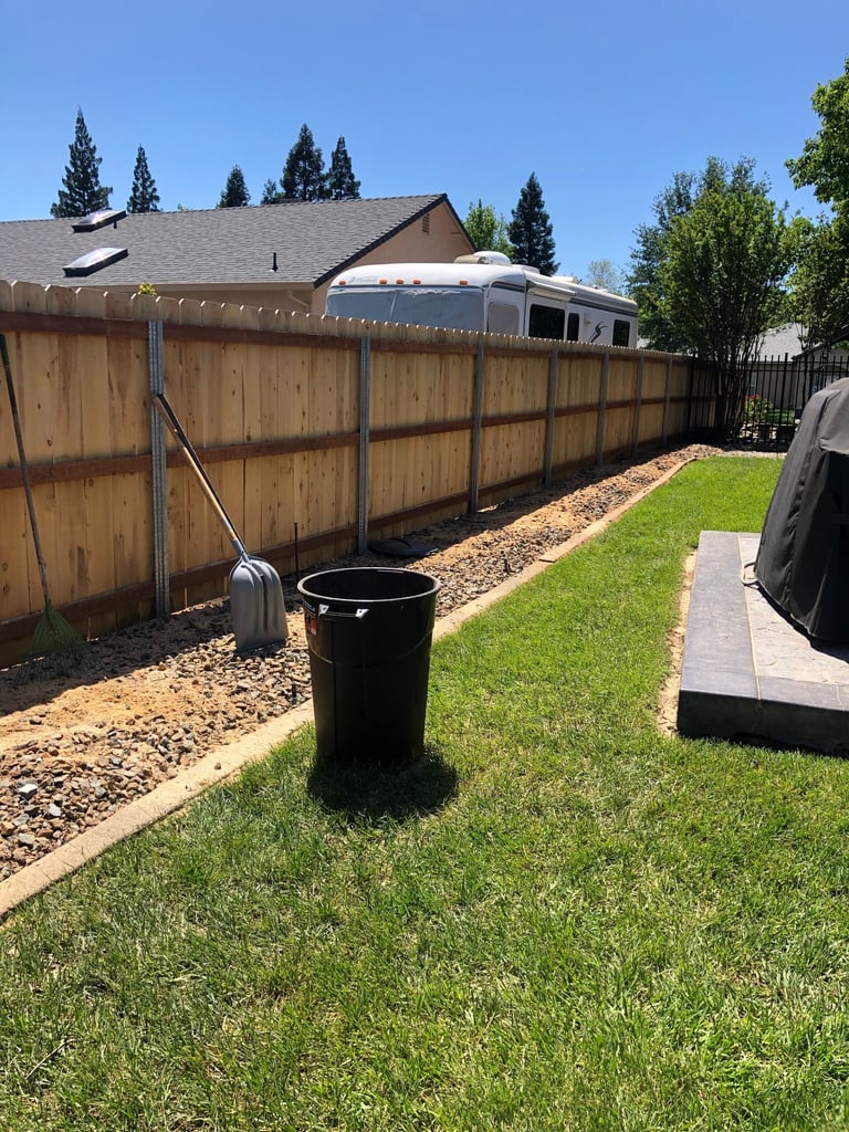 a grassy backyard with cleared area and trash can near fence line 