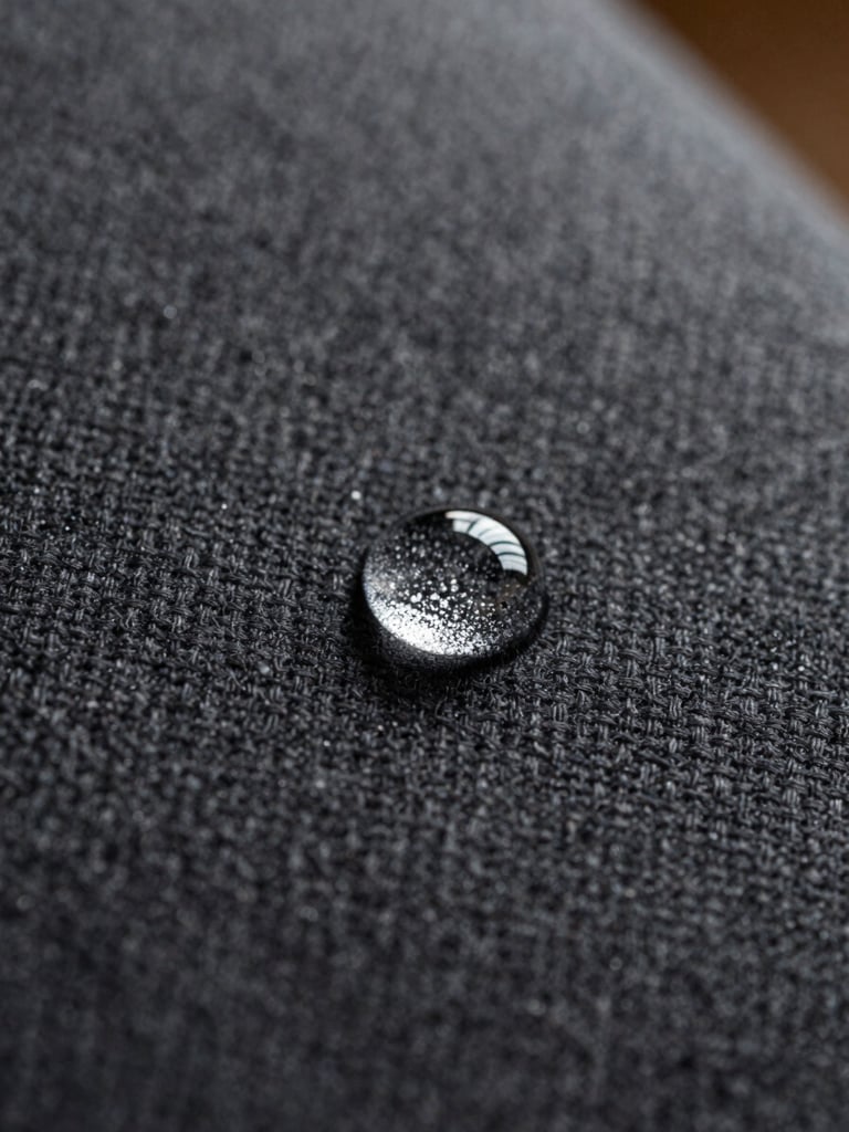 Extreme close-up, overhead angle — a single water droplet beading on dark charcoal sofa fabric in bright natural north-facing daylight, the bead perfectly spherical against the weave, fabric texture sharp and unaltered
