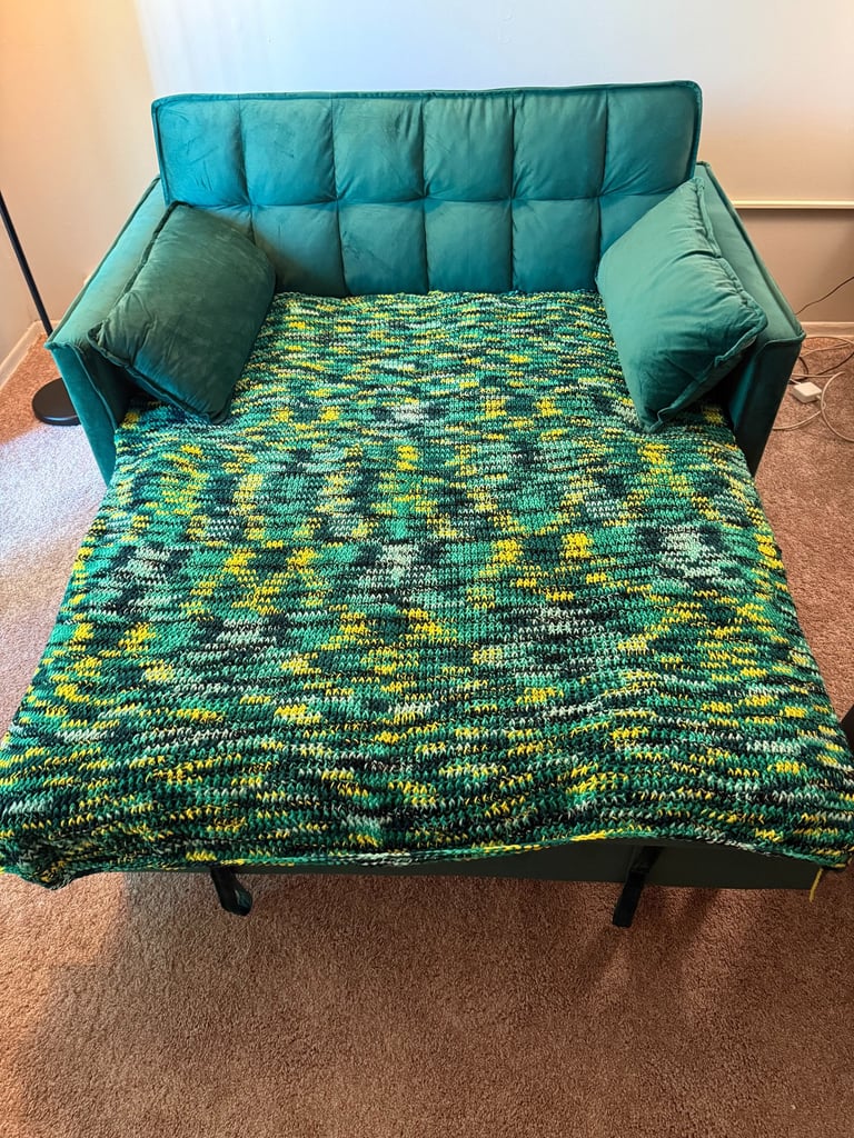 A green velvet convertible sofa bed covered with a handmade green and yellow variegated crochet blanket.