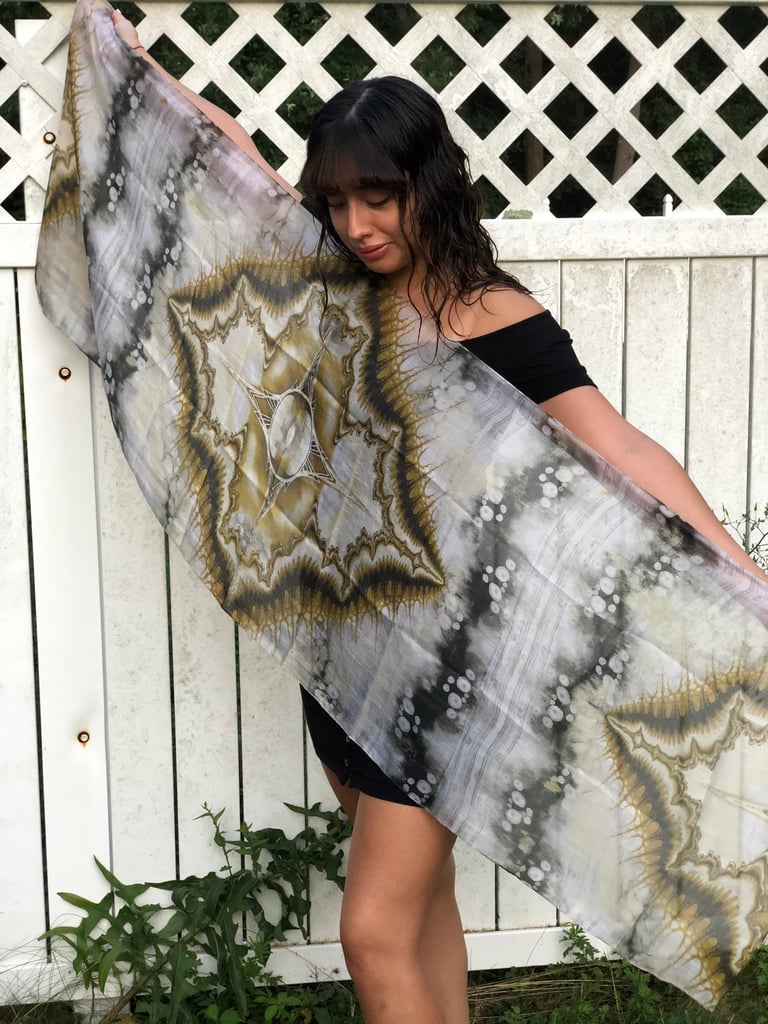 A woman holding up a lightweight silk scarf featuring an intricate hand-dyed earth tone mandala pattern.