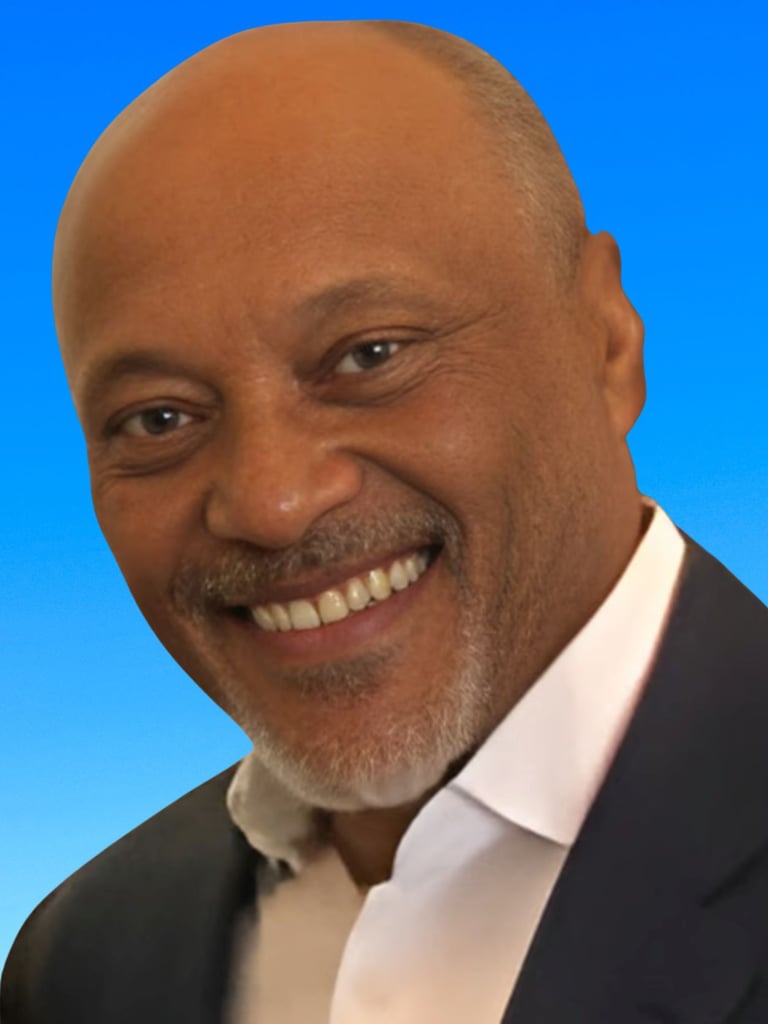 A professional portrait of a smiling bald man with a graying goatee wearing a blazer against a blue background.