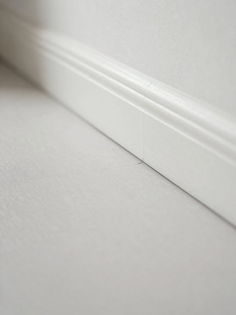 Straight-on close-up of a perfectly flush vinyl floor joint meeting a clean white baseboard, bright clinical daylight, dust-free surface, architectural lines.
