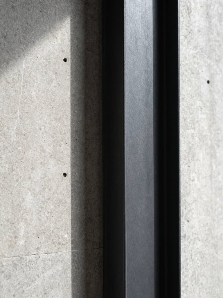 Macro photograph of a raw concrete wall meeting a clean black steel structural column under harsh directional sunlight, highlighting textures.