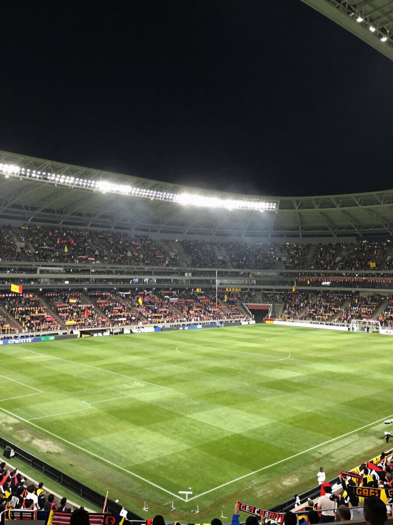 Panoramic wide shot of a packed Colombian football stadium at night, green pitch glowing under floodlights, crowd in the far stands holding up scarves and flags, atmospheric haze above the field, electric anticipation before kickoff