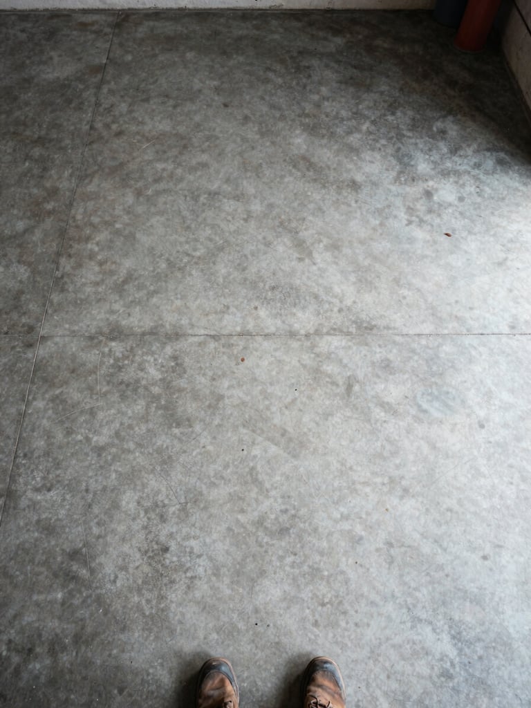 Overhead wide-angle shot in natural daylight — a completely empty garage floor after a full cleanout, bare concrete visible, no clutter remaining, a single pair of work boots at the bottom edge of the frame hinting at the crew that just finished