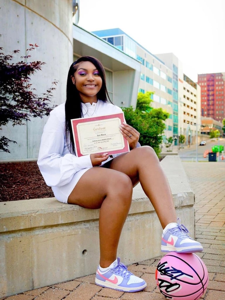 A smiling graduate in a white dress holds a certificate next to a signed pink basketball.