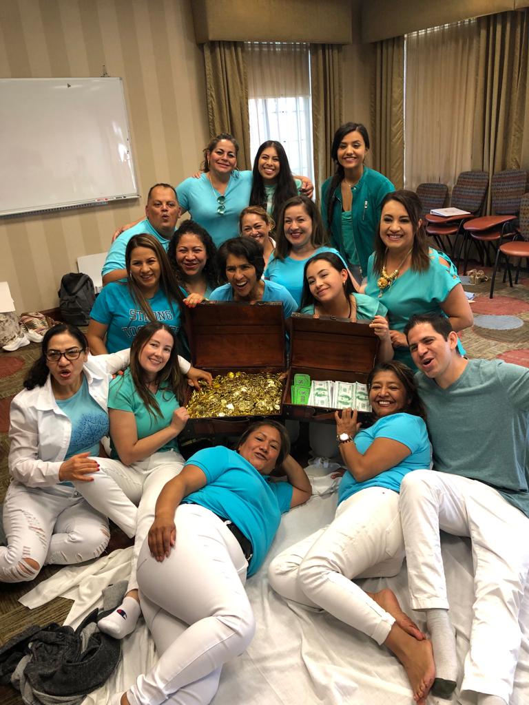A cheerful group of 18 women and 2 men, dressed in blue and white, pose together around a treasure chest overflowing with
