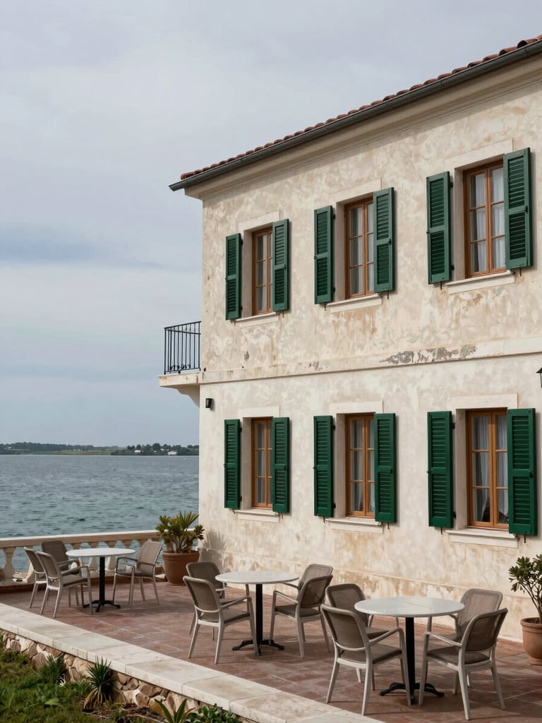 Environmental wide shot of a two-storey waterfront hotel facade in overcast daylight, terrace with empty chairs overlooking the Adriatic sea, weathered pale render and dark-green shutters, lagoon visible in background, no people, available light only