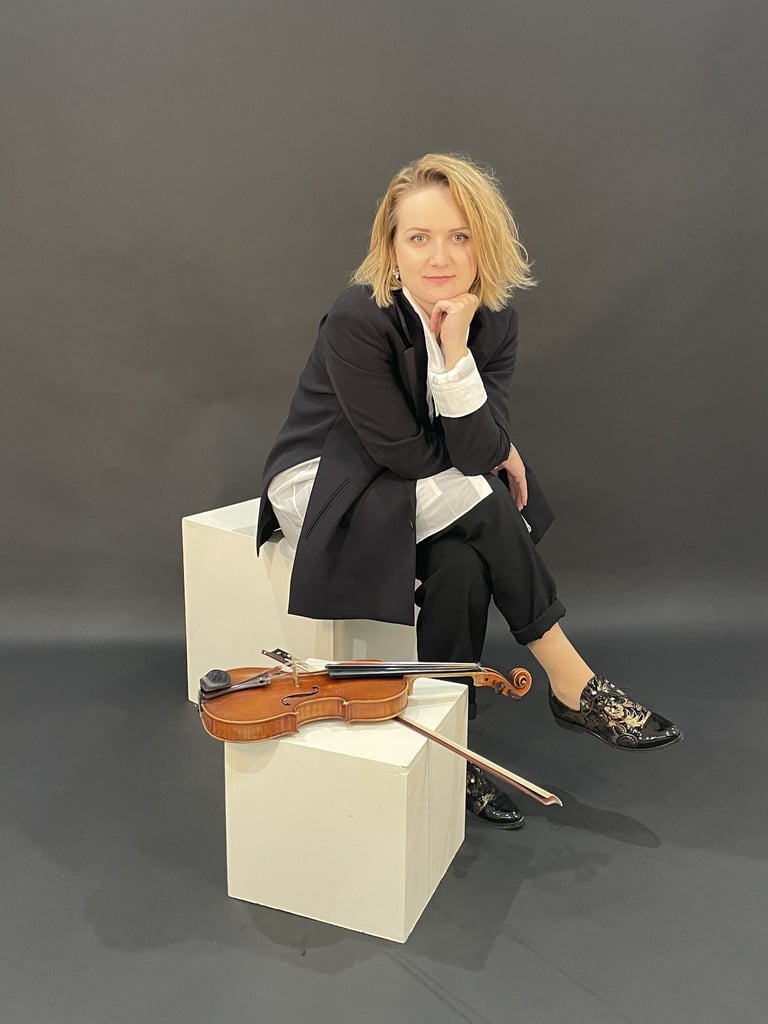 Violinist Žydrė sitting in a studio, leaning forward with hand under chin, violin resting beside her