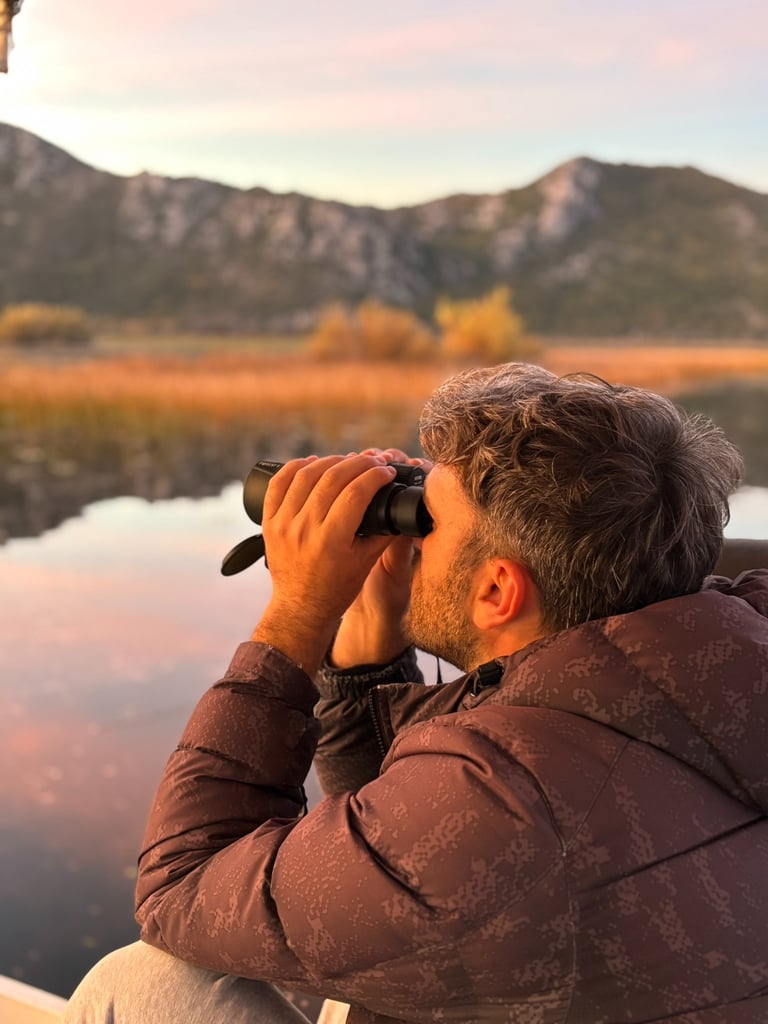 A man uses binoculars for birdwatching over a lake with scenic mountain views at sunset.