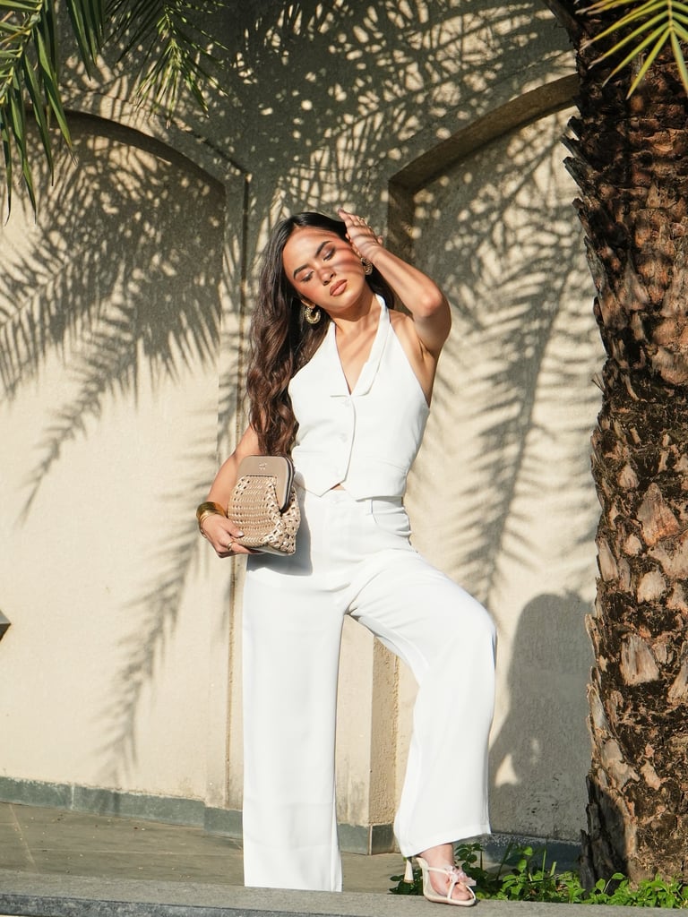 Woman posing in a white halter vest and wide-leg trousers holding a straw clutch bag under palm trees.