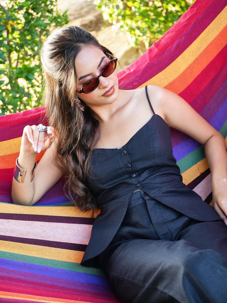 A stylish woman in a black linen outfit and sunglasses relaxing on a colorful striped hammock outdoors.