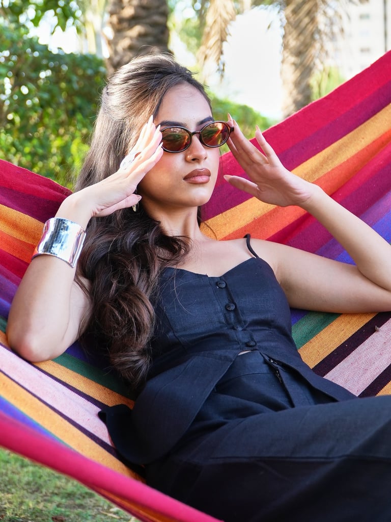 A fashionable woman in a black outfit and sunglasses relaxes in a colorful striped hammock outdoors.