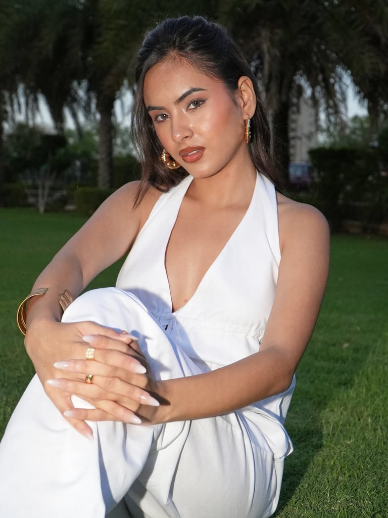 A woman posing in a white halter top and gold jewelry on a lush green lawn.