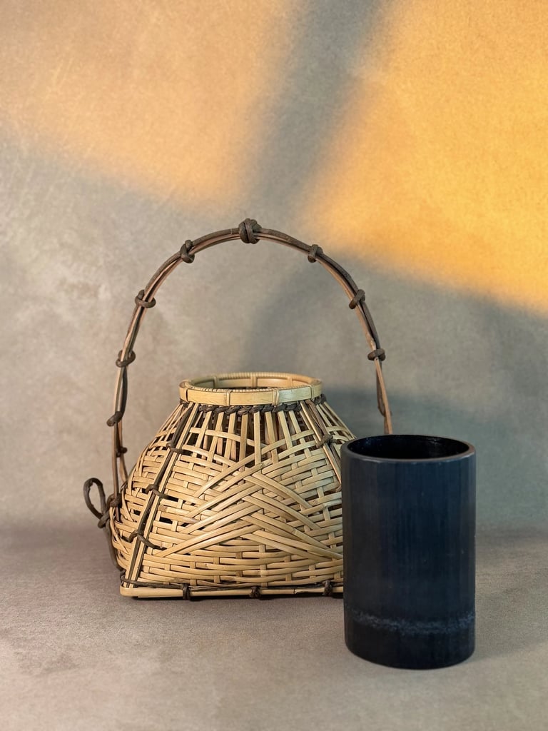 Handcrafted woven bamboo basket with handle next to a black ceramic vase in warm light.