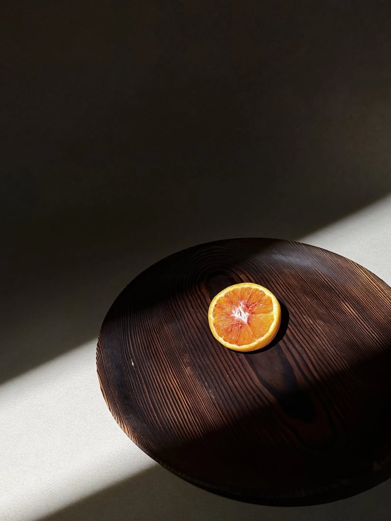 A single blood orange slice centered on a dark wooden platter under dramatic natural sunlight.