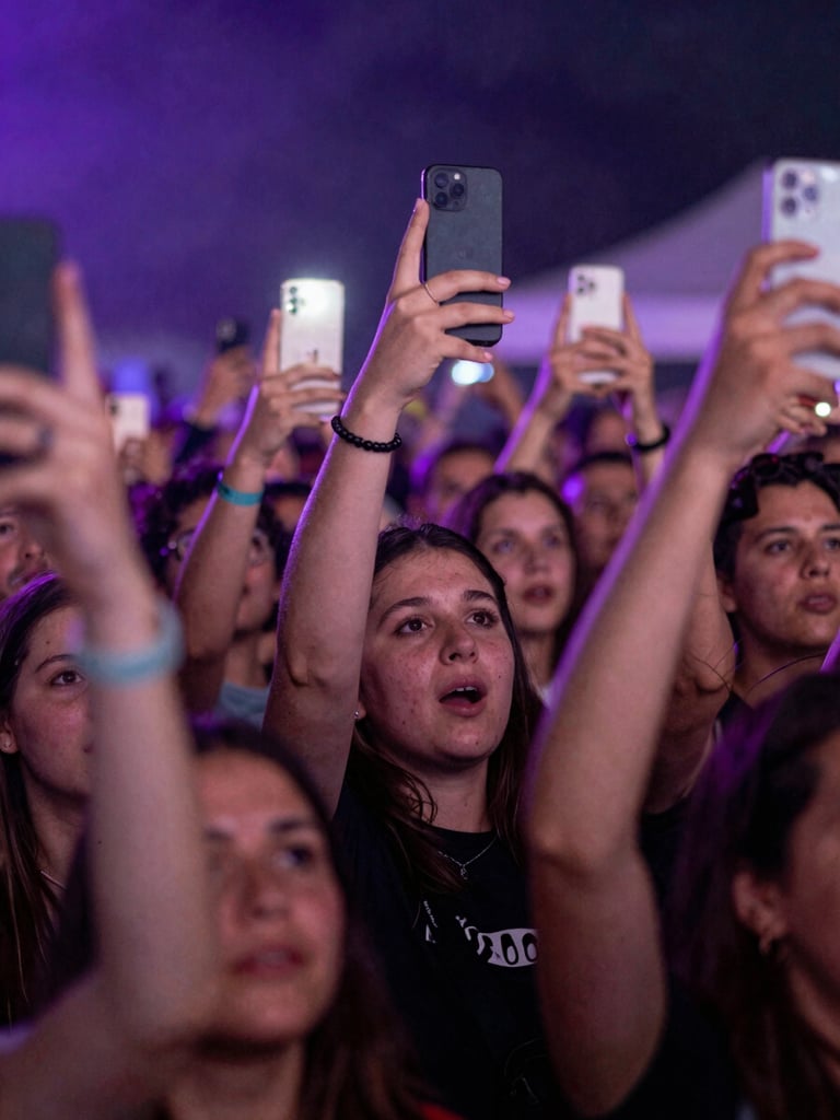 Close-up portrait frame of a crowd mid-cheer at a festival, faces lit simultaneously by stage wash in electric violet and the glow of phone screens held aloft, physical and digital worlds visible in the same moment, high-contrast lighting, tight emotional framing