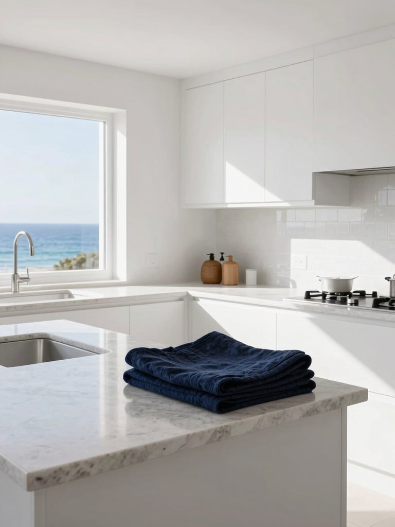 A bright, sunlit coastal kitchen with clean white cabinets, natural stone countertops reflecting morning light, and neatly folded linens on the island.