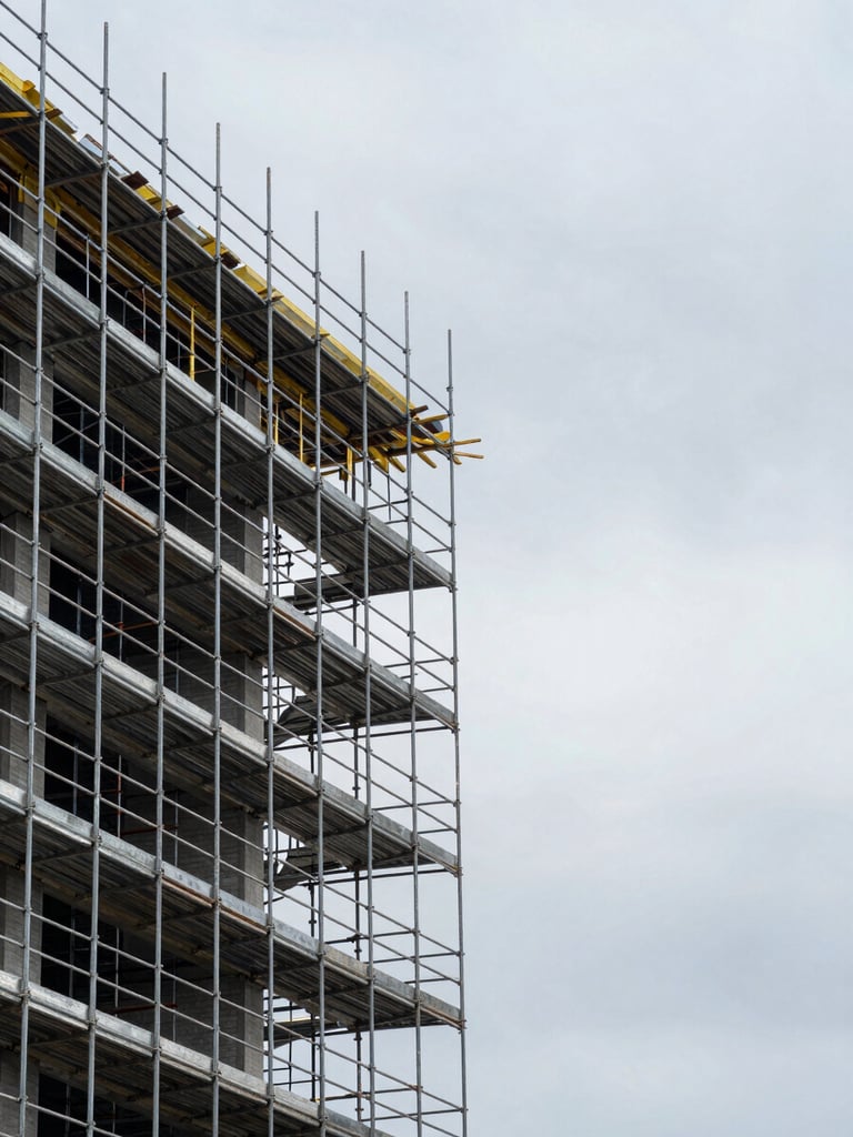 Wide shot of an active construction site showing an assembled ringlock scaffold structure on a mid-rise building facade — overcast daylight, galvanized steel catching soft sky reflection, no workers visible, structure occupying left two-thirds of the frame with open sky to the right