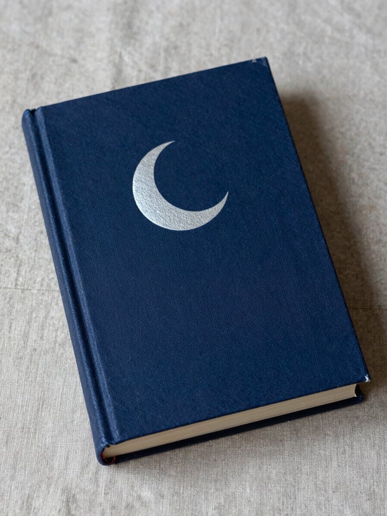 Close portrait of a hardbound book lying on aged linen fabric, cover facing up — deep indigo cloth binding with a pale silver crescent moon embossed at center, soft candlelight raking across the texture of the cover from lower left, cool shadow on the right third, no text visible, intimate cabinet-of-curiosities framing