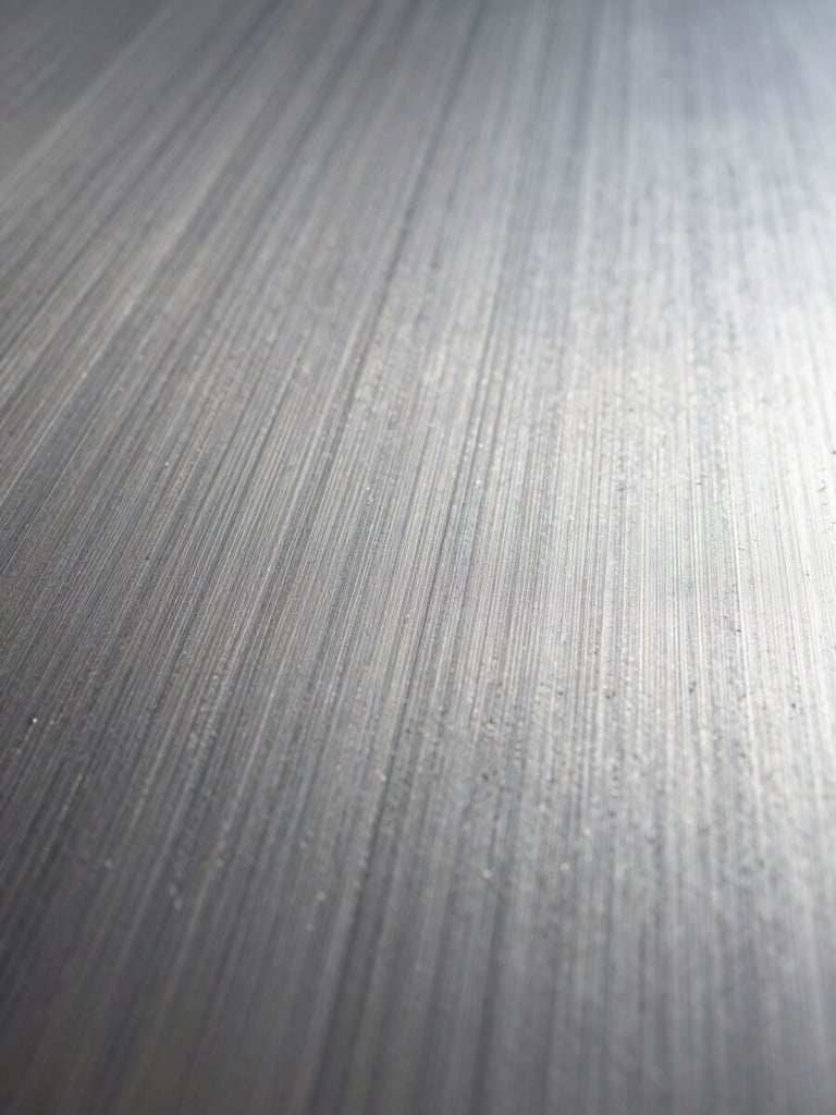Extreme close-up of a brushed steel surface, studio strobe lighting raking across the grain at a low angle, tight crop revealing micro-texture and parallel tool marks, no subject other than the material itself, wide negative space toward the right edge