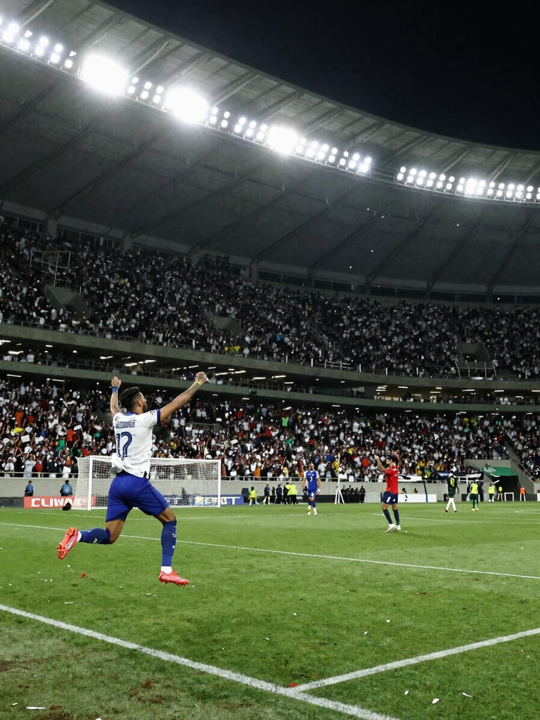 Wide environmental shot, Brazilian football stadium packed with fans, a striker mid-air celebrating a goal in the left foreground, stadium floodlights blazing overhead, green pitch stretching to the right, crowd confetti mid-air, natural stadium lighting, intense and immediate