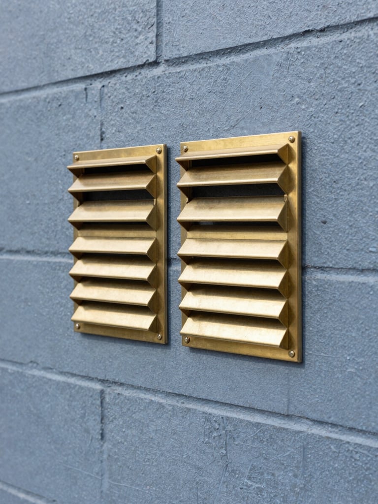 A close-up of clean, modern brass subfloor vents neatly installed into a heritage double-brick wall, bright daylight, sharp commercial photography.