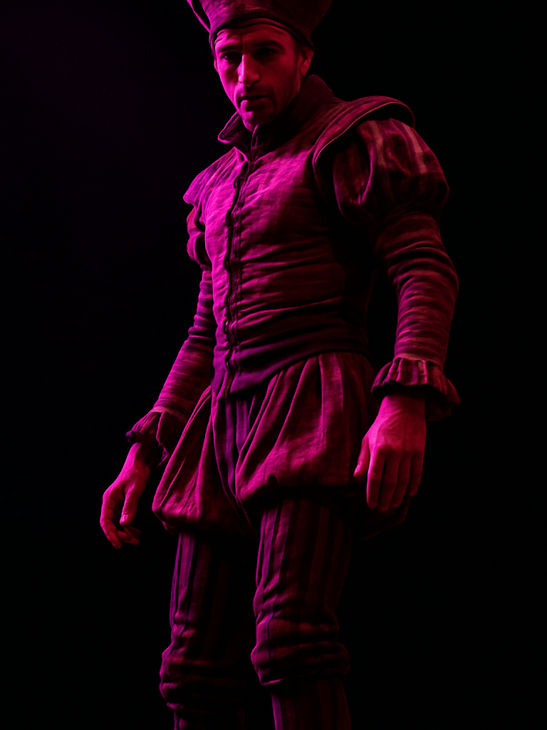 Tight vertical frame, performance lighting from stage left — a zanquero in full costume locks eyes with someone in the crowd, upper body and stilts visible, magenta gel light cutting across the figure against deep black background